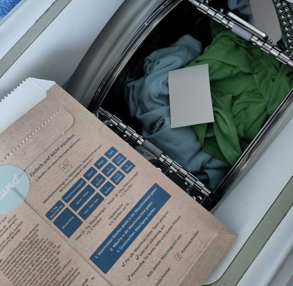 Toplader-Waschmaschine gefüllt mit hellblau und grünen Kleidungsstücken, obenauf liegt ein rechteckiger heller Waschstreifen 